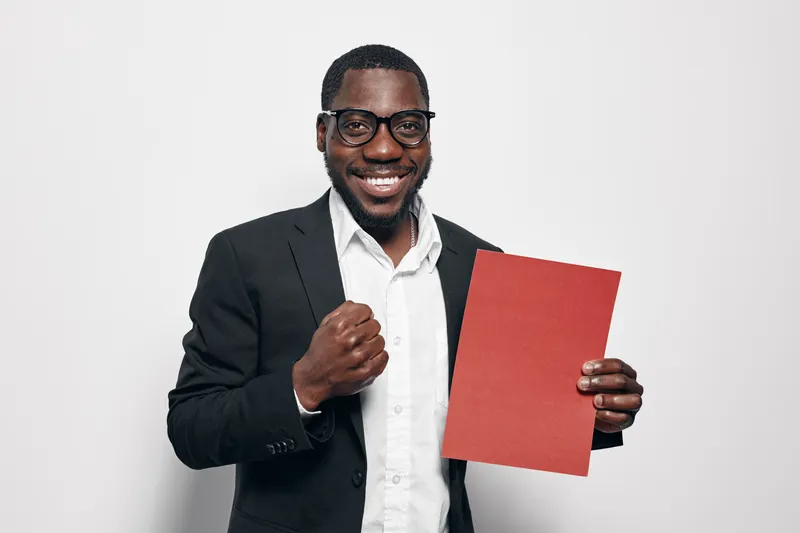 African American man with dark skin wearing glasses and black suit holding red folder and smiling confidently on white background for business success and achievement.