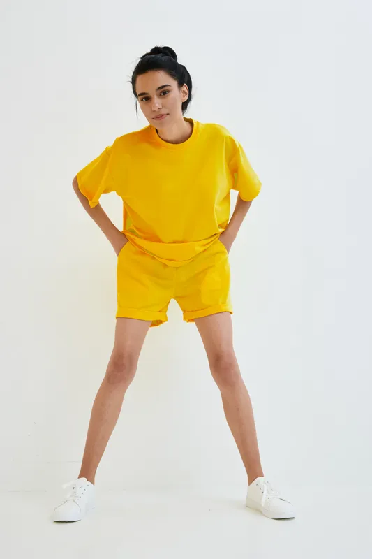 young woman in yellow casual outfit stands confidently with hands on hips against white background, showcasing relaxed style and modern fashion trends in bright color.