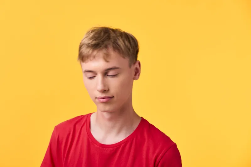 Calm young man in a red shirt with closed eyes rests against a bright yellow background, conveying serenity, focus, and relaxation in a simple lifestyle composition for wellness and mindfulness themes