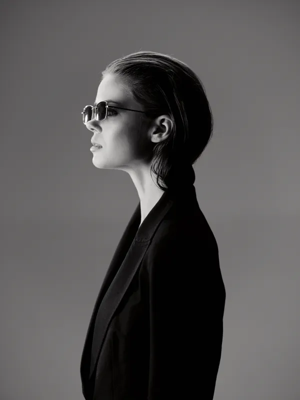 Black and white portrait of a stylish woman wearing sunglasses and dark blazer with slicked-back hair in profile view.