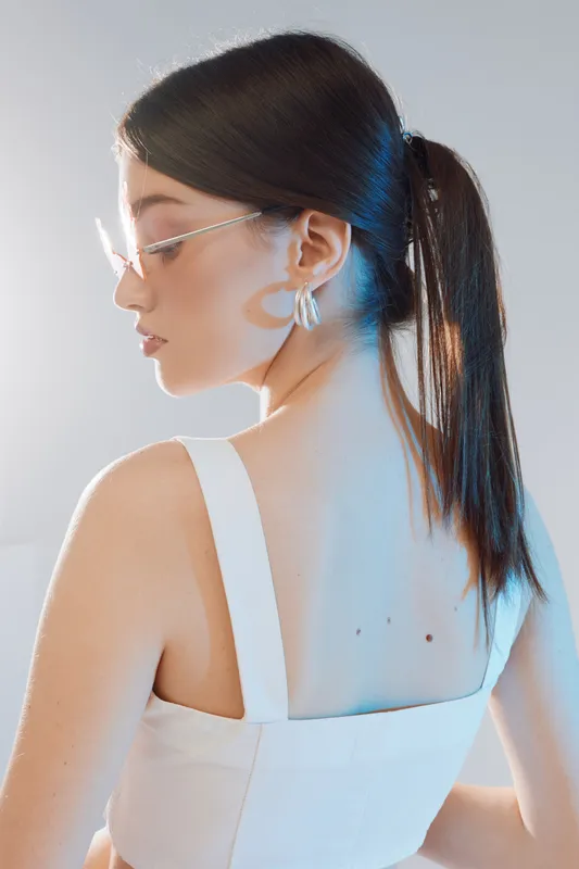 Young woman with long dark hair in a white top and glasses poses sideways against a light background. Concept of beauty, fashion, elegance, and modern style.