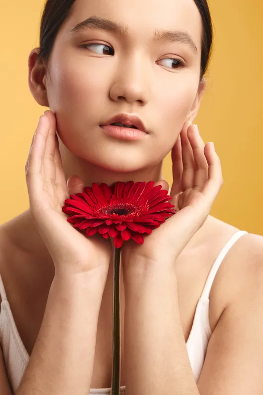 Asian woman is pictured in a studio portrait, holding a red flower near her face, serene expression, warm lighting, and graceful pose conveying calm beauty.