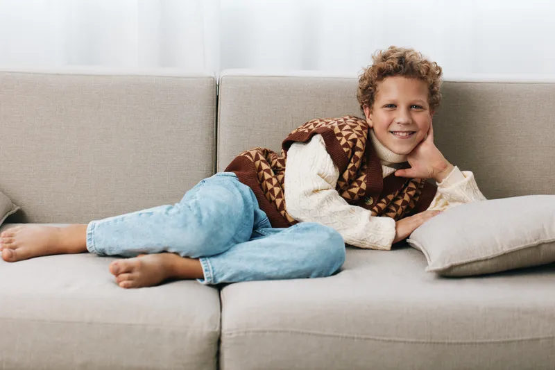 A young boy relaxing on a light sofa, lying on his side with a warm knitted vest and denim jeans, smiling softly while resting his head on a cushion.