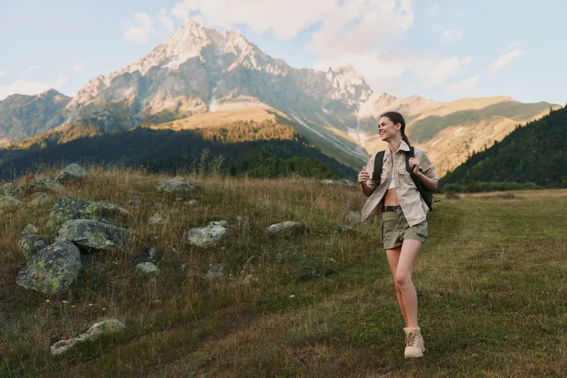 Young woman with a backpack hikes across a sunlit alpine meadow beneath towering mountains, her casual jacket fluttering as she explores the trail with a sense of freedom.