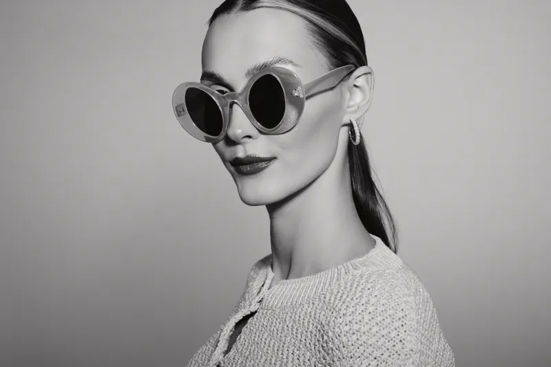 Portrait of a fair skinned Caucasian woman wearing large round sunglasses and a knitted sweater against a plain studio background.