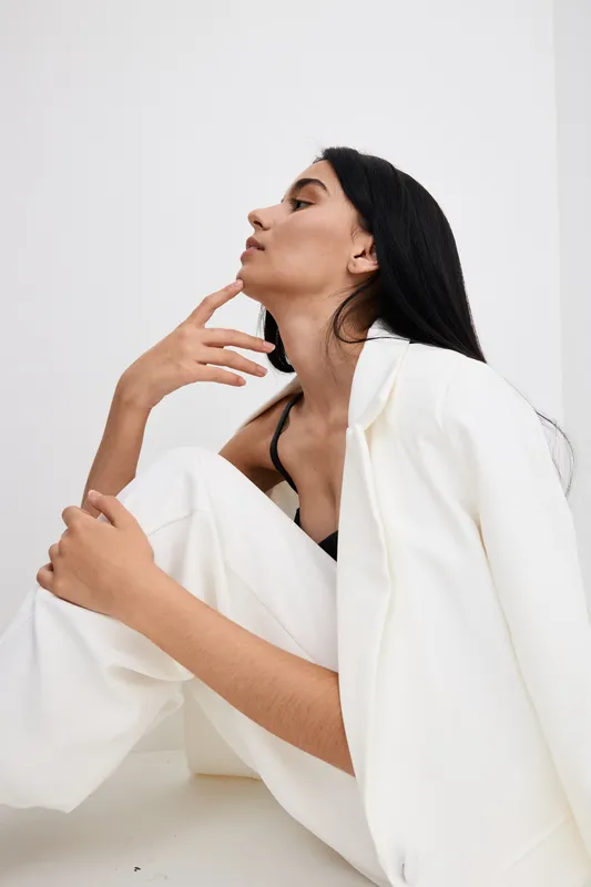 tall woman with long black hair wearing white suit sitting thoughtfully against plain white background, stylish, minimalistic, contemplative mood
