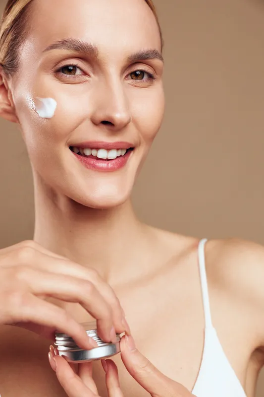 Smiling woman in studio portrait applying face cream from jar, skincare routine for moisturizing beauty, spa cosmetics treatment, gentle care, soft light, beige background