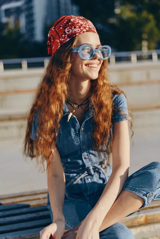 Lifestyle woman in modern boho-western denim street style with red bandana and round glasses, smiling outdoors under natural sunlight, captured in film color tones.