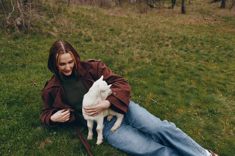 Woman relaxing on grass outdoors, cuddling a white goat kid. Casual clothing, brown jacket, green sweater. Warm, cozy mood highlights affection, bonding, countryside, animal companionship, nature,