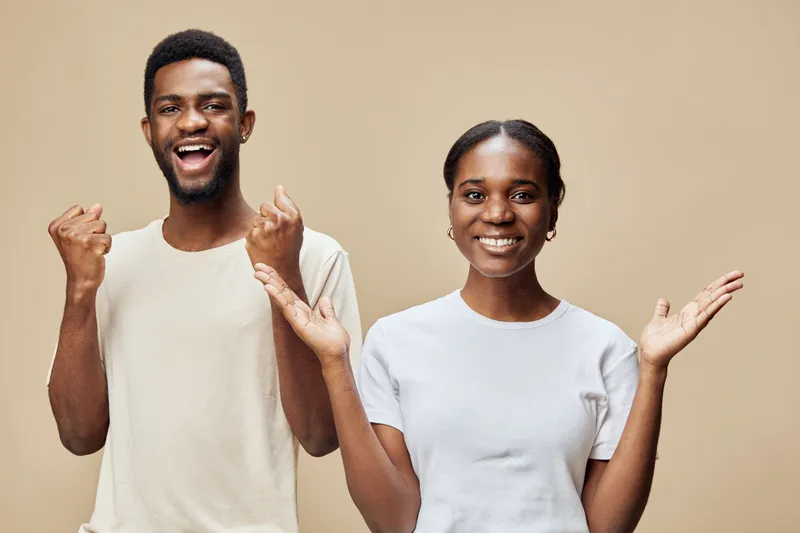 Excited young African American couple celebrating success with joyful expressions against a warm beige background, wearing casual light colored clothing