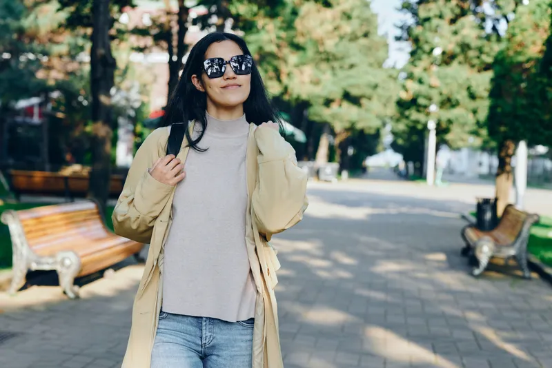 Woman wearing a long beige coat and sunglasses strolling through a sunlit park, casual fashion vibe, daytime street scene, relaxed pose amid greenery benches and warm light