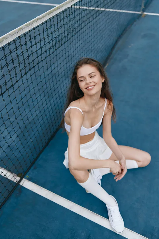 Cheerful female tennis player in white sportswear, seated on a court, exuding confidence and joy, perfect for sports and healthy lifestyle concepts