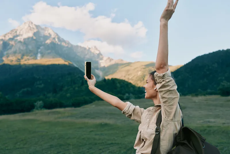 Smiling woman in a beige jacket taking a selfie in a green meadow with mountains in the background, embodying outdoor lifestyle, travel and exploration adventure