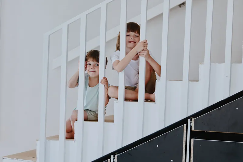 Two playful children sitting on a staircase, smiling joyfully. The bright, minimalistic interior enhances their cheerful expressions, creating a lively atmosphere.