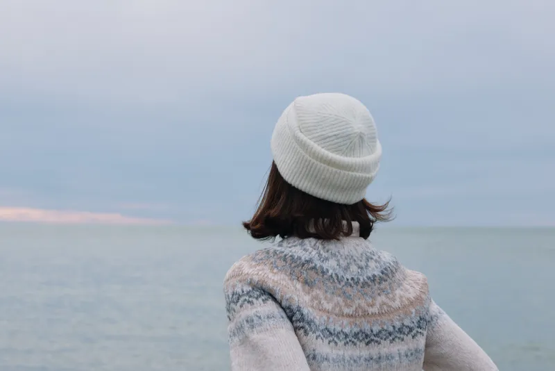 Woman with back view wearing a white beanie and a patterned sweater by the sea, calm horizon, soft light, and minimalist fashion mood perfect for lifestyle and travel stock.
