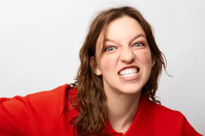 Angry young woman with brown hair wearing red sweater shows teeth and grimaces against white background. Concept of frustration, emotion, and intense facial expression in studio.