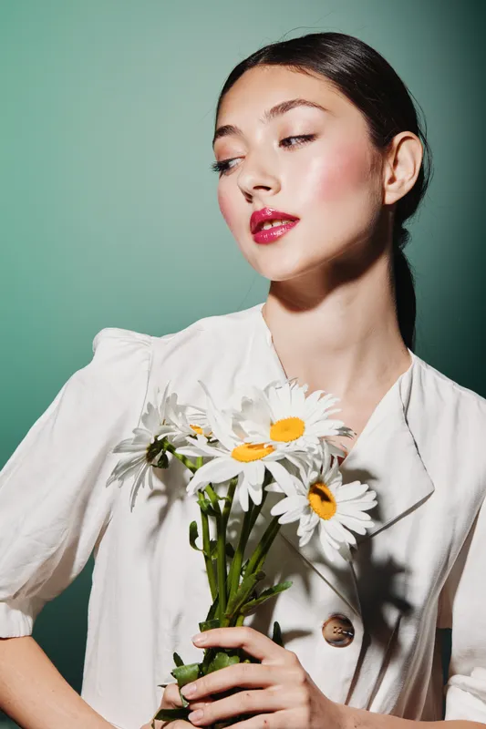 Young woman with pink glossy lips holding white daisies in a white blazer looks aside on a green background. Creative beauty and fashion concept with feminine elegance.