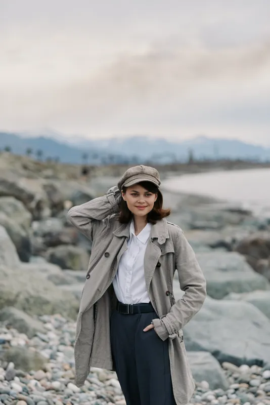 Young woman in casual fall outfit stands on rocky beach with mountains in the background. She wears a hat and coat, smiling with hand on head, enjoying a cool outdoor autumn day.