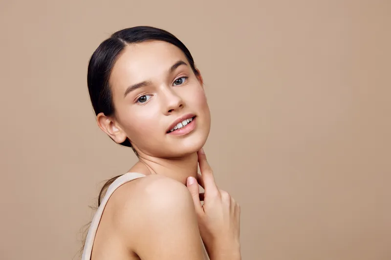 Young woman with natural beauty gazes playfully at the camera, showcasing skincare routine and healthy lifestyle against a soft beige background