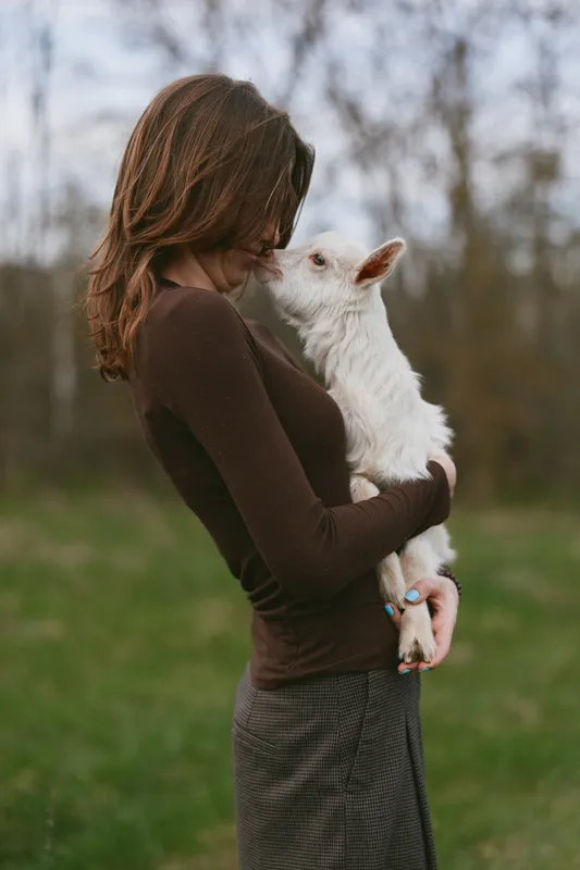A young woman with long brown hair holds and kisses a small white baby goat in a green outdoor setting, capturing a tender lifestyle moment of connection and care.