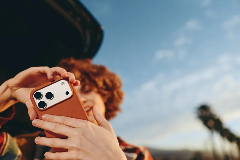 Woman lifestyle outdoors holding smartphone mobile hands gripping device technology communication, curly hair woman in background, sunny sky backdrop, blurred pedestrian far away, summer travel mood