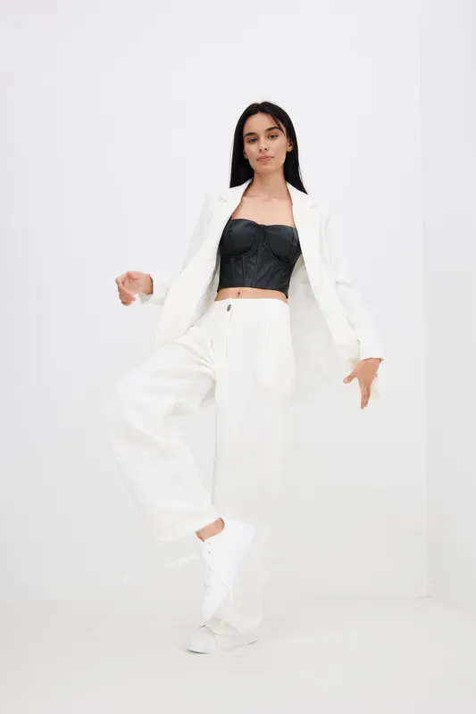 Young woman in white suit and black top confidently posing in studio with minimalistic white background, expressing modern fashion and casual elegance.