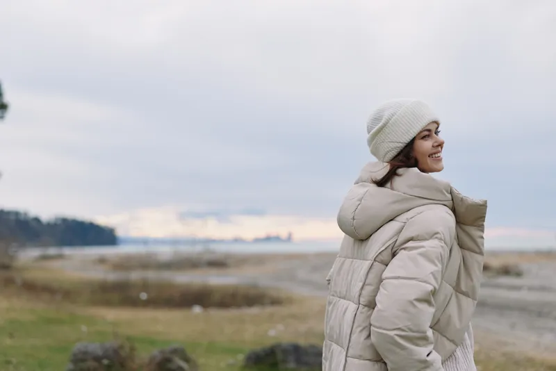 Woman in a beige puffer coat and beanie gazes toward a windy seaside landscape with a calm horizon, creating a serene outdoor fashion portrait.