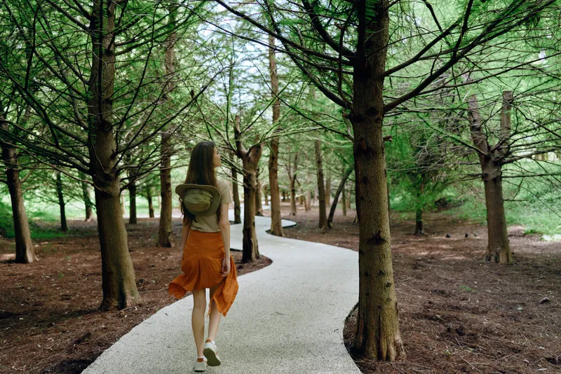 Woman walking on a winding path through a green forest with backpack, orange skirt and sneakers. Solitary traveler on a gravel trail among tall trees for peaceful nature hike.