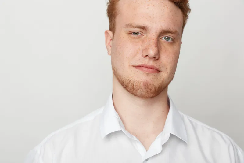 Confident young man with red hair and beard wearing white shirt looks at camera with slight smile on plain light background. Concept of casual style, portrait, and natural expression.