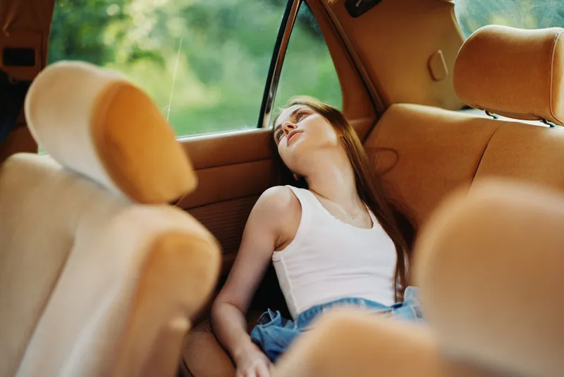 A young woman resting in a vintage car, showing a serene expression, surrounded by warm brown upholstery, evoking a sense of nostalgia and tranquility in nature.