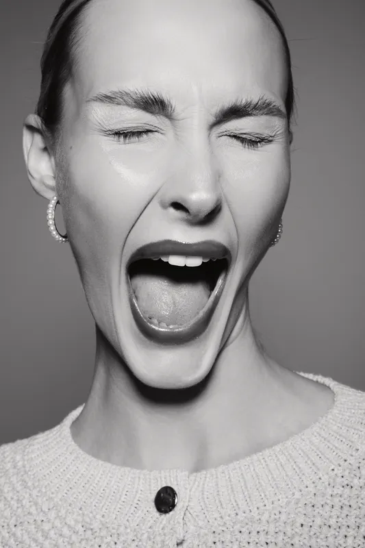 White woman with light skin and closed eyes screams or shouts in emotional black and white close-up portrait.
