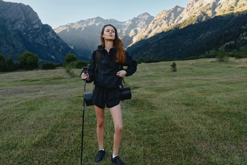 A confident female photographer stands in a wide valley with dramatic mountains in the background, gripping a camera and tripod, ready to capture outdoor scenery with focused energy.