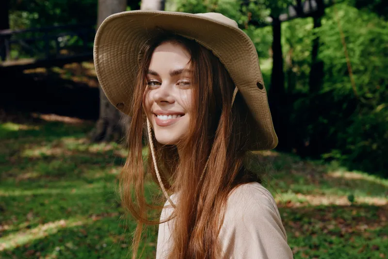 Woman smile hat portrait nature outdoors: young woman wearing a wide brim sunhat, smiling in a sunlit park with long hair and casual style for summer lifestyle and outdoor portrait.
