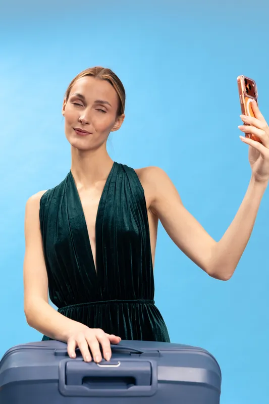 Young woman in elegant dress takes selfie with smartphone while holding suitcase handle. Blue background studio portrait. Concept for travel, fashion, lifestyle, and vacation preparation.