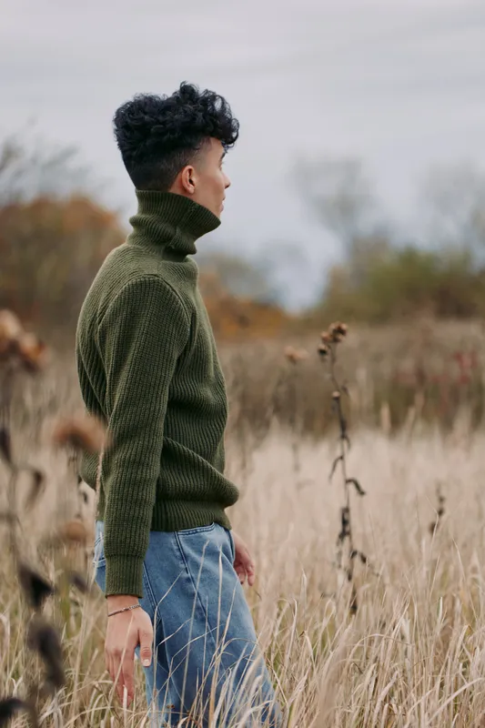 A young person in a textured green sweater stands in tall autumn grass, captured in a candid, authentic moment that emphasizes genuine presence and reliable everyday style.