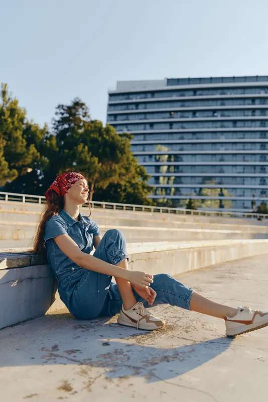 relaxed woman sitting outdoors in casual denim outfit and white sneakers with red bandana, enjoying calm sunny day near urban park and modern building background