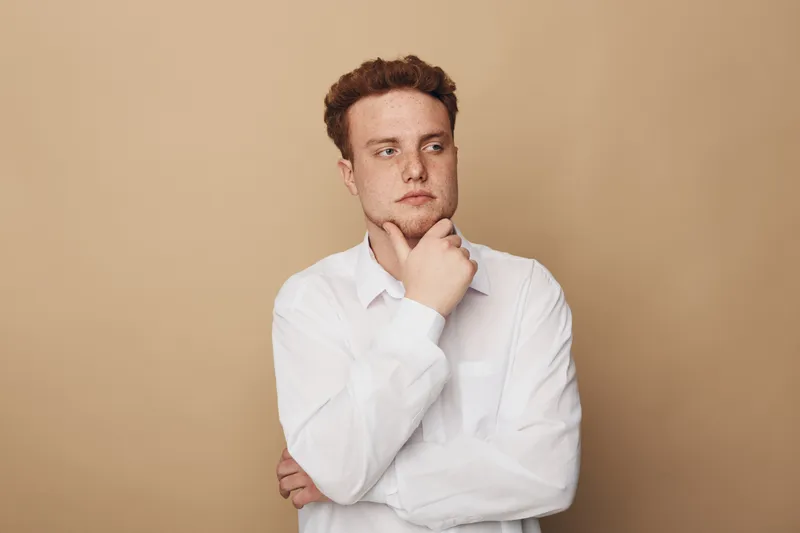 Thoughtful young man in white shirt looks away with hand on chin against beige background. Concept of contemplation, decision making, and introspection in casual style portrait.