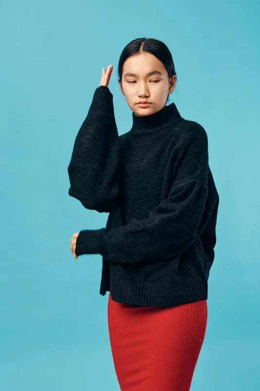 Young woman posing with eyes closed wearing black turtleneck sweater and red skirt on blue background. Fashion style, casual outfit, calm mood, studio portrait with soft lighting.