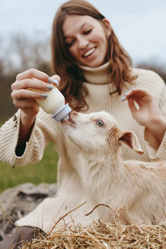 A woman outdoors feeds a young goat with a bottle, smiling as the goat drinks. Warm natural tones, soft breeze, rustic setting capturing a tender lifestyle moment