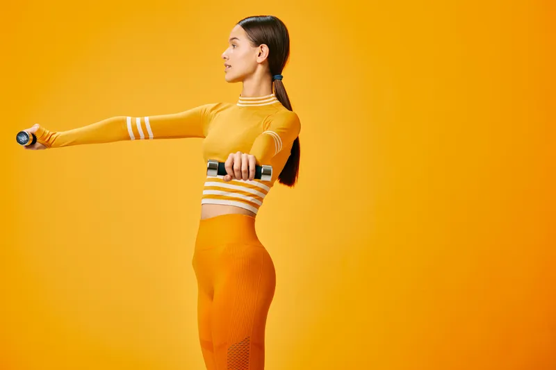 Woman fitness athlete in bright orange activewear extends her arms with dumbbells in a vibrant studio, showcasing athletic form, balance and dynamic sport training.