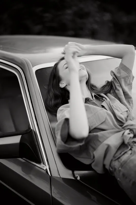 Black and white portrait of a young woman enjoying a carefree moment by a vintage car, showcasing relaxed emotions and a timeless style.