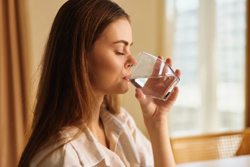 Woman drinking water from a glass, portrait closeup of a young woman at home in the morning. Relaxed female hydrating in a robe, natural light, wellness and everyday self care routine.