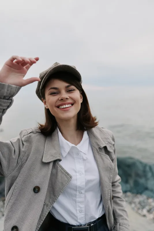 Smiling young woman in casual coat and hat stands near rocky seaside, enjoying fresh air and natural light. Concept of happiness, outdoor lifestyle, and autumn fashion.
