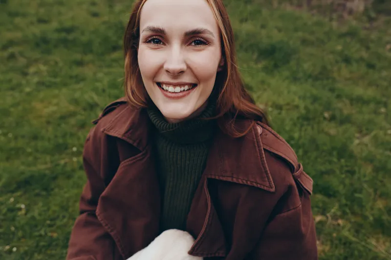 Young woman with red hair smiles warmly while holding a small white baby goat outdoors on a green grass field during a casual lifestyle moment.