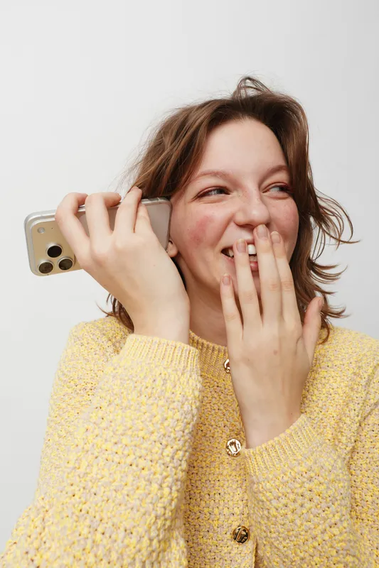 Smiling young woman in yellow sweater holds smartphone near ear, laughing and covering mouth with hand. Concept of happiness, communication, and casual lifestyle.