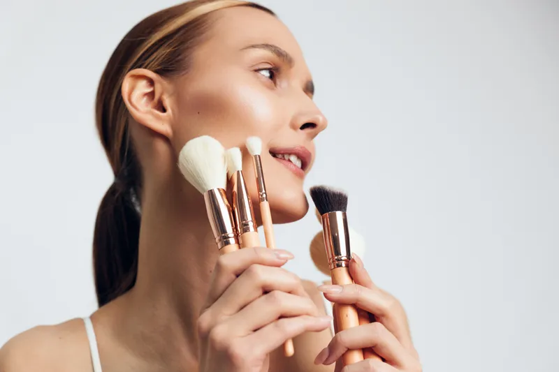 A close-up of a young woman smiling softly as she holds makeup brushes near her face, ready to apply powder, showcasing a calm beauty routine and self-care.