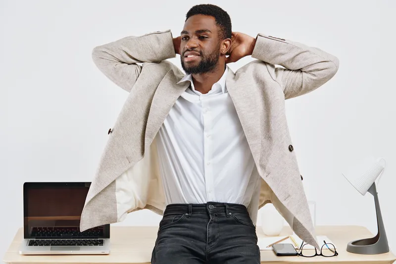 Confident young African American man in casual blazer and white shirt standing relaxed in modern office environment, smiling. Studio portrait with clean background, professional appearance, and positi