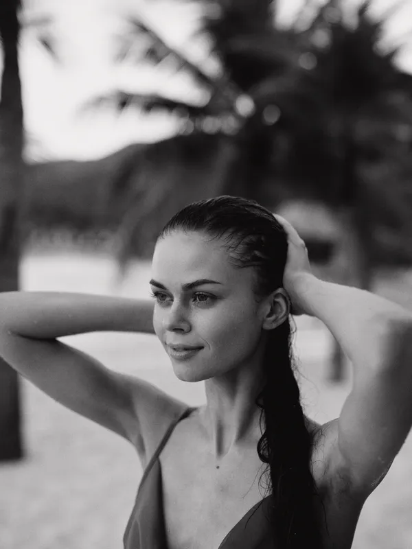 Black and white portrait of a young woman at the beach, arms raised to touch her hair, calm expression, soft lighting, timeless and elegant moment.