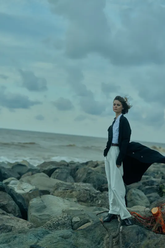 Woman on a rocky shoreline in windy weather, flowing coat and white pants, seascape backdrop with moody clouds and ocean horizon