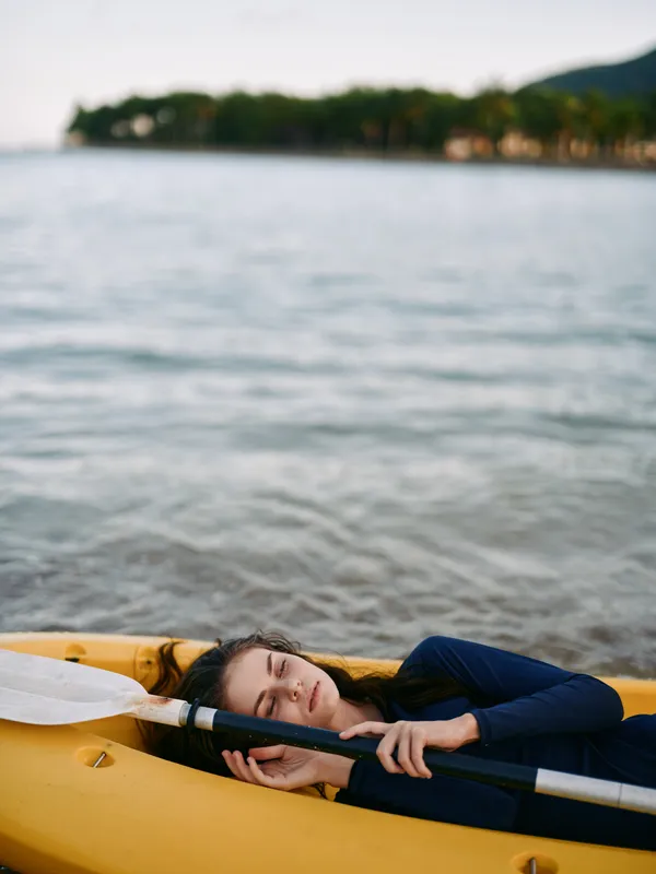 A person resting in a yellow kayak on calm water, close shot of face and paddle, capturing a peaceful recreational boating moment under soft light and open sky.
